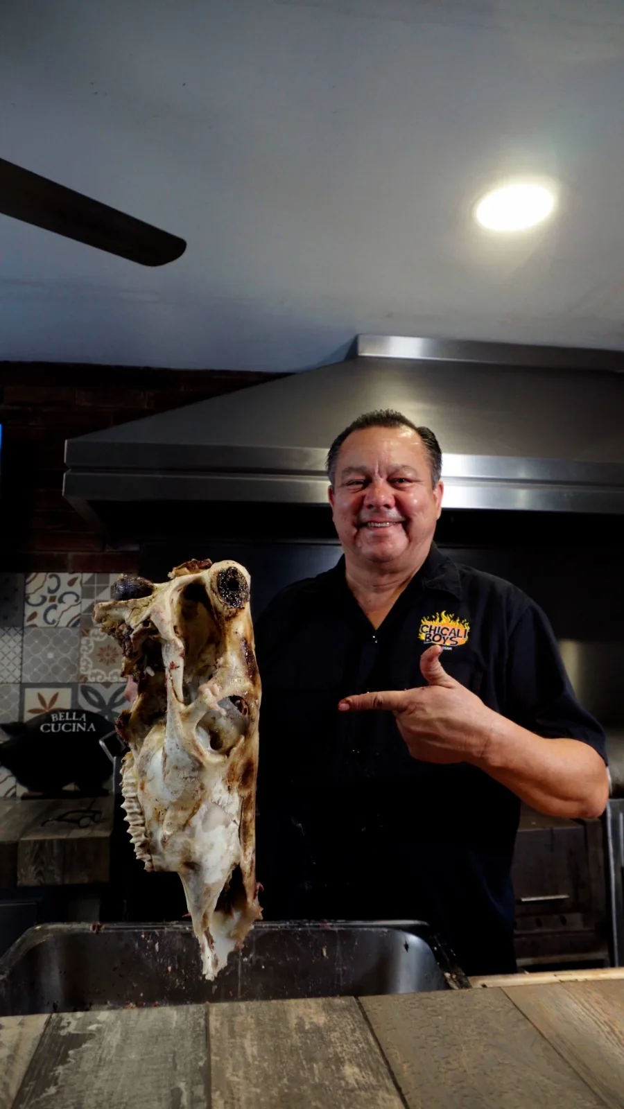 Integrante de Chicali Boys sonríe en la cocina mientras muestra el hueso de una cabeza de res ya cocida.