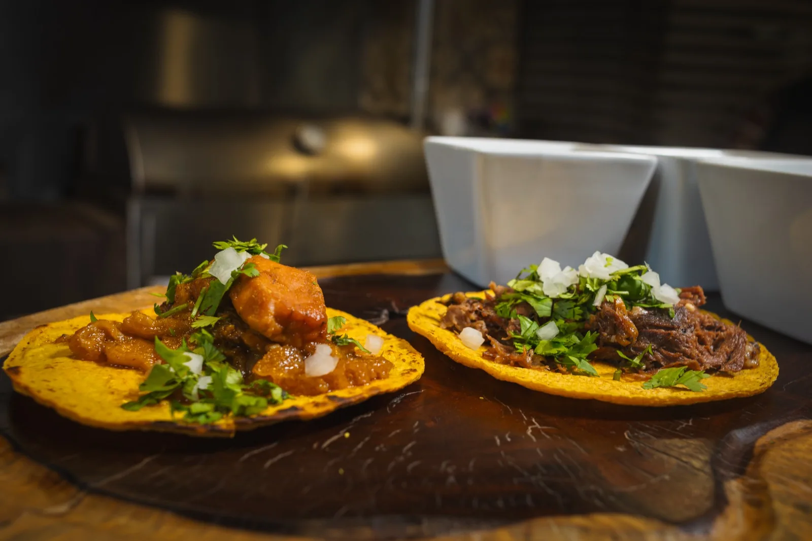 Dos tacos con cebolla y cilantro sobre una tabla de madera, con tazones blancos al fondo.