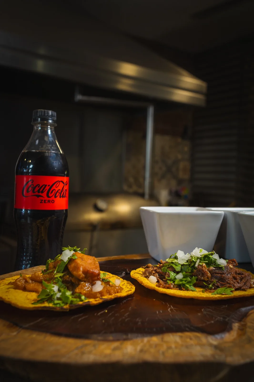 Dos tacos sobre una tabla de madera con un refresco de botella al fondo.